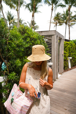 Woman in a straw hat and white dress holding a phone and pink bag on a wooden path with greenery and palm trees in the background.
