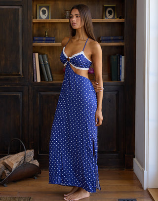 A woman modeling a blue and white polka dot maxi dress with a cutout waist, standing in front of a dark wood bookshelf.