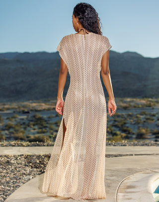 Woman in a beige knit dress standing on a wooden platform with a mountainous landscape in the background.