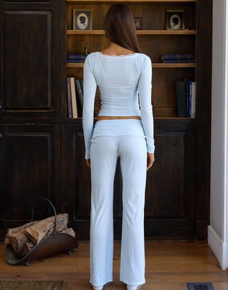 A woman stands with her back to the camera wearing a light blue ribbed long-sleeve crop top and matching high-waisted flare pants, positioned in front of a dark wood bookshelf.