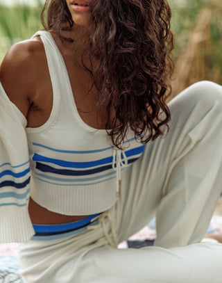 Woman wearing a white tank top with blue stripes and beige pants sitting outdoors.