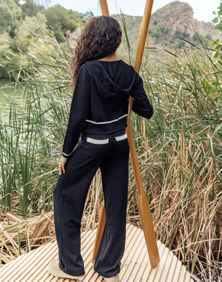 Person wearing a black outfit with white accents standing on a wooden platform surrounded by reeds and water.
