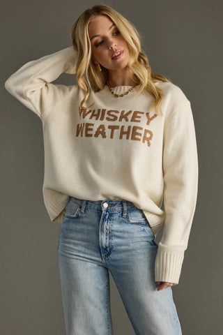 Woman wearing a cream sweater with 'Whiskey Weather' text and blue jeans on a gray background