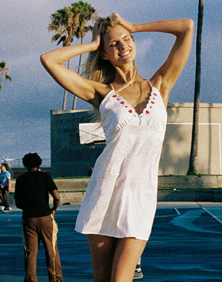 A woman posing on an outdoor basketball court wearing a white sleeveless mini dress with red heart details on the neckline.